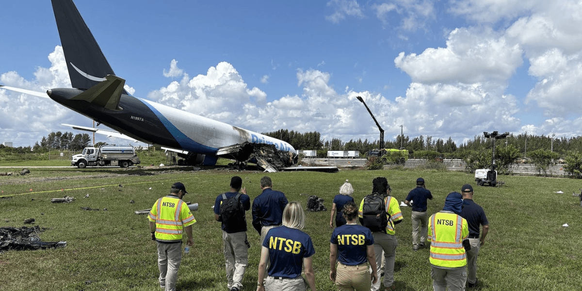 NTSB investigators approaching a plane crash site in a grassy area with clouds above