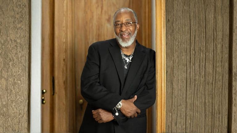 A smiling man with a gray beard, wearing glasses and a black suit jacket, stands with arms crossed. He leans against a wooden door frame, exuding warmth.