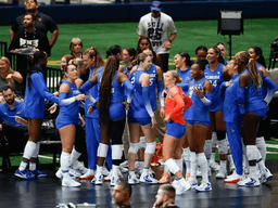 A volleyball team in blue uniforms huddles together, celebrating on the court.