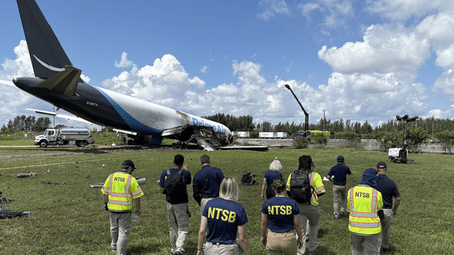 NTSB investigators approaching a plane crash site in a grassy area with clouds above