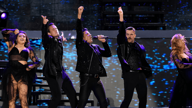 Five performers in matching outfits energetically singing on stage with bright lights