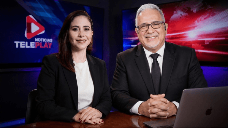 Two news presenters in formal attire sitting at a desk with a laptop and studio background