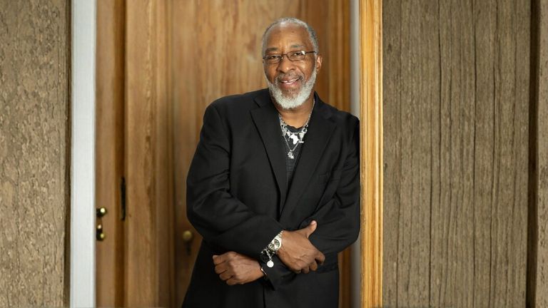 A smiling man with a gray beard, wearing glasses and a black suit jacket, stands with arms crossed. He leans against a wooden door frame, exuding warmth.