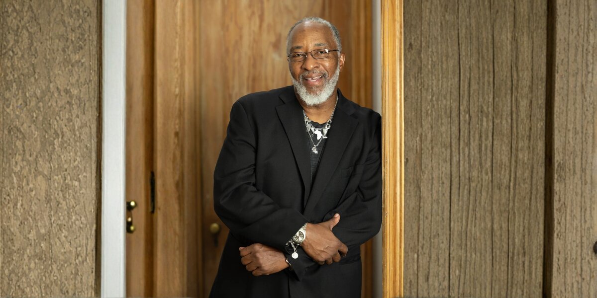 A smiling man with a gray beard, wearing glasses and a black suit jacket, stands with arms crossed. He leans against a wooden door frame, exuding warmth.