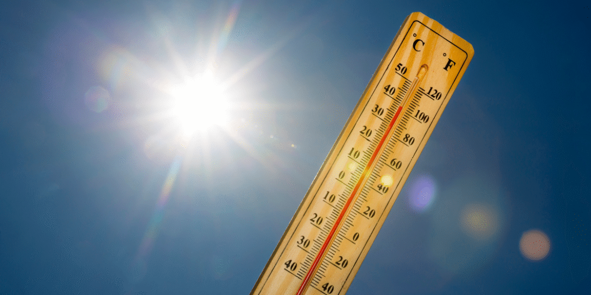 Wooden thermometer showing high temperatures beneath a bright sun in a clear blue sky.