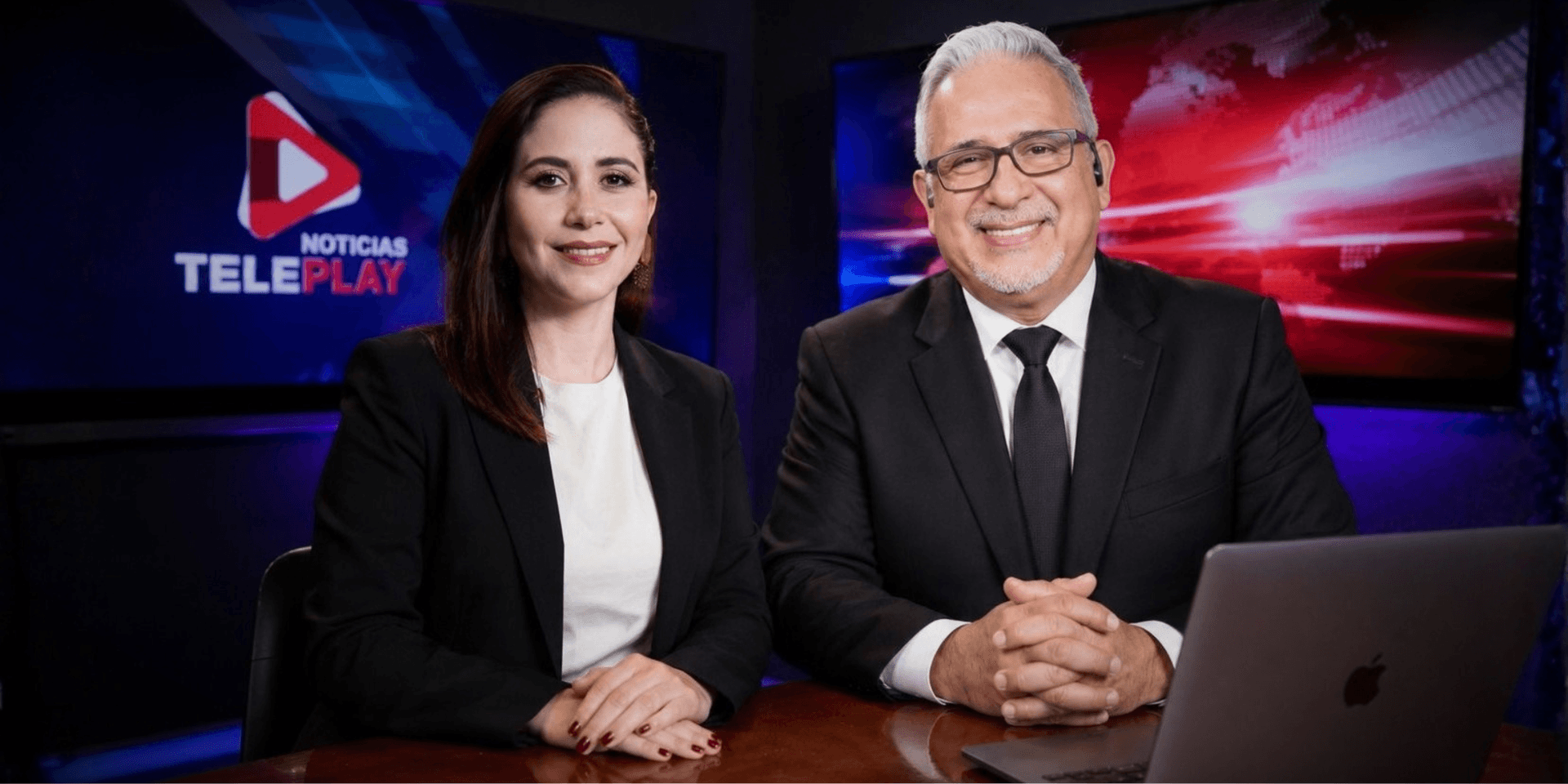 Two news presenters in formal attire sitting at a desk with a laptop and studio background