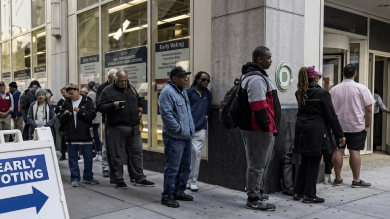 Voters wait in line outside an early voting location with a directional sign in the foreground.